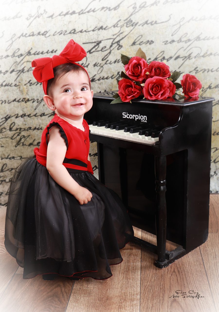 Bebé con moño rojo junto a piano de juguete, Erica Cruz