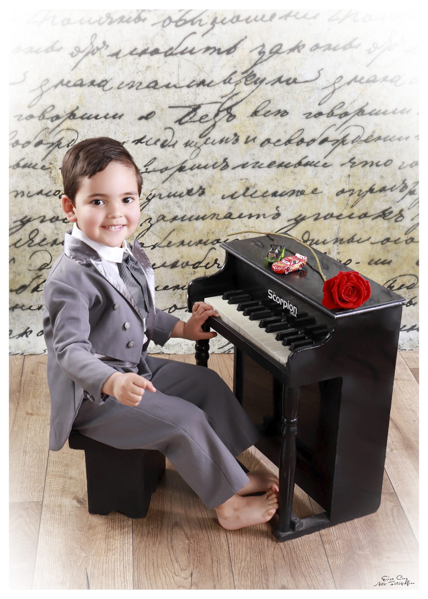 Niño con traje gris junto a piano de juguete, Erica Cruz