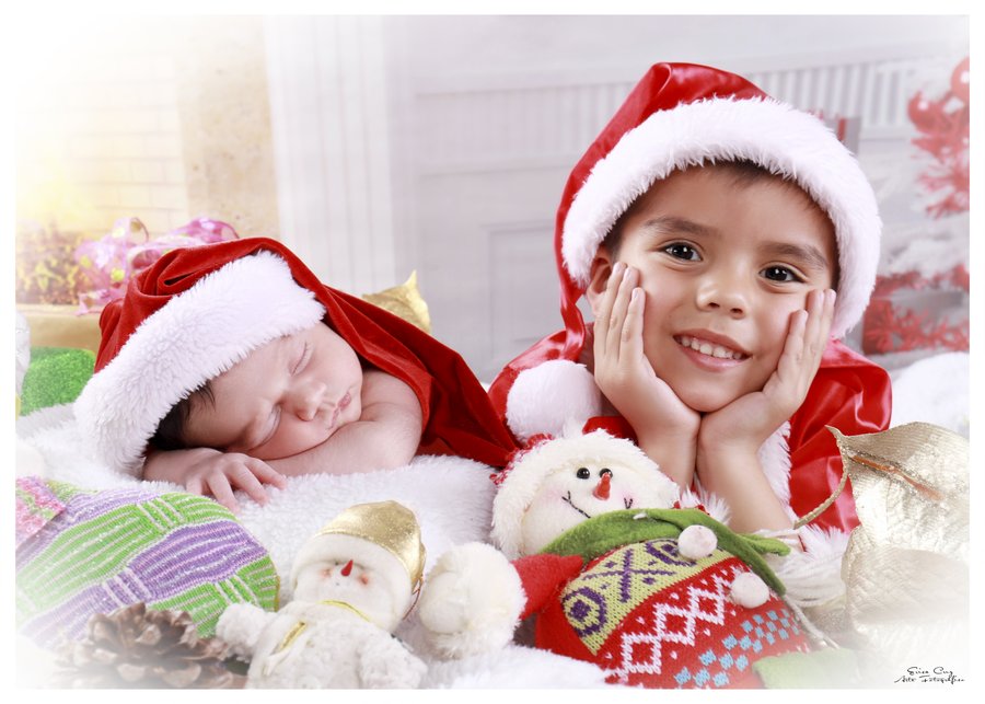 Hermano mayor junto a su hermana recién nacida dormida, ambos con gorro navideño, Erica Cruz