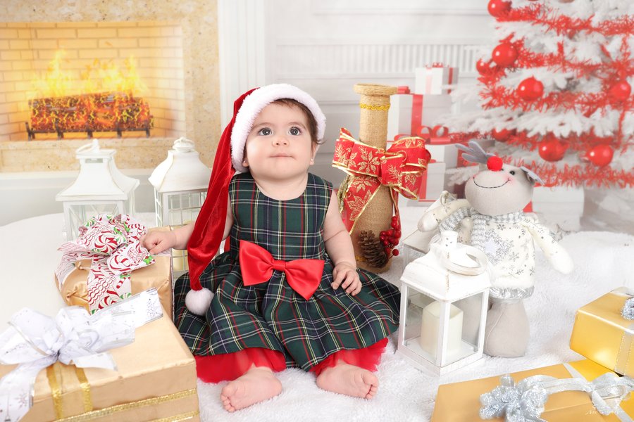 Bebé con vestido escocés y gorro navideño junto a la chimenea, Erica Cruz