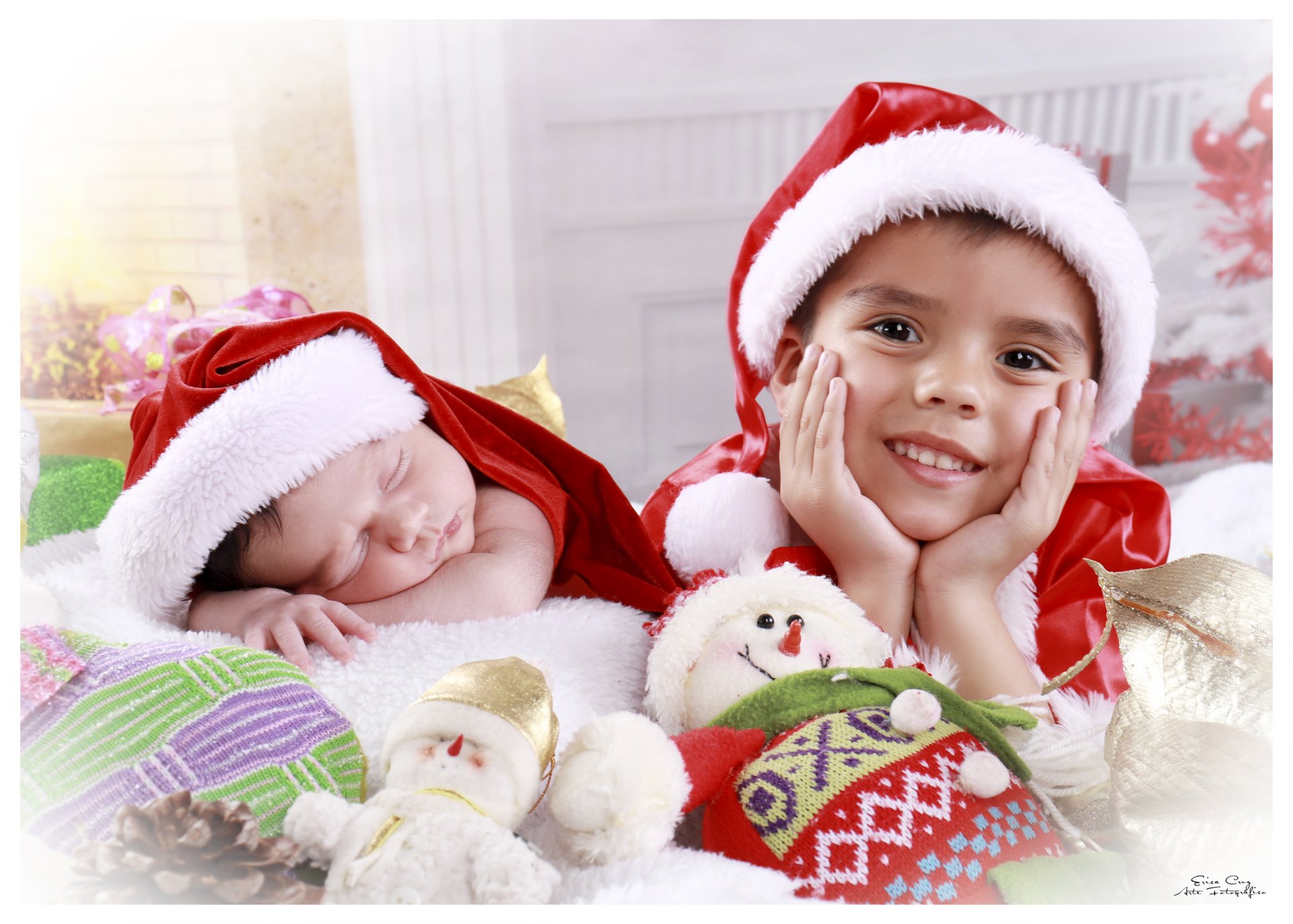 Hermano mayor sonriendo junto a su hermana recién nacida dormida, ambos con gorro navideño, Erica Cruz Santiago